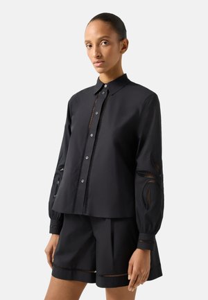 Kvinde iført en sort knap-op skjorte med udskårne blomsterpatter på ærmerne og matchende sorte shorts mod en ensfarvet lys baggrund.