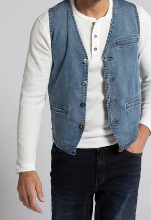 Mężczyzna w białej koszuli henley z długim rękawem, niebieskiej dżinsowej kamizelce na guziki i ciemnych dżinsach, z jedną ręką w kieszeni na jednolitym tle.