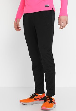 Persona con pantaloni sportivi neri Nike, scarpe da corsa Nike arancione brillante e una maglia a maniche lunghe rosa, in piedi su un pavimento e uno sfondo bianchi.