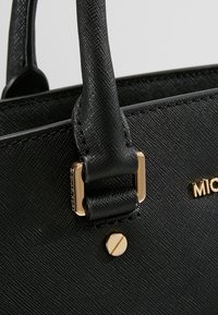 Gros plan sur la poignée d'un sac à main en cuir noir texturé, attachée avec une ferrure rectangulaire dorée et un détail visible de vis dorée.