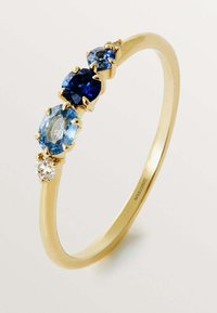 Anillo de oro con tres piedras preciosas: un gran zafiro azul, un topacio azul claro en forma ovalada y un pequeño diamante transparente, todas engastadas en garras.