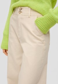 Beżowe spodnie z zapięciem na dwa guziki, kieszeniami bocznymi i gładką fakturą, połączone z grubym zielonym swetrem z dzianiny.