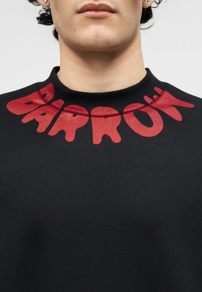 Κοντινό πλάνο ενός ατόμου που φοράει μαύρο πουκάμισο με μεγάλα κόκκινα γράμματα που γράφουν "ΠΑΠΑΓΑΛΟΣ" γύρω από το γιακά.