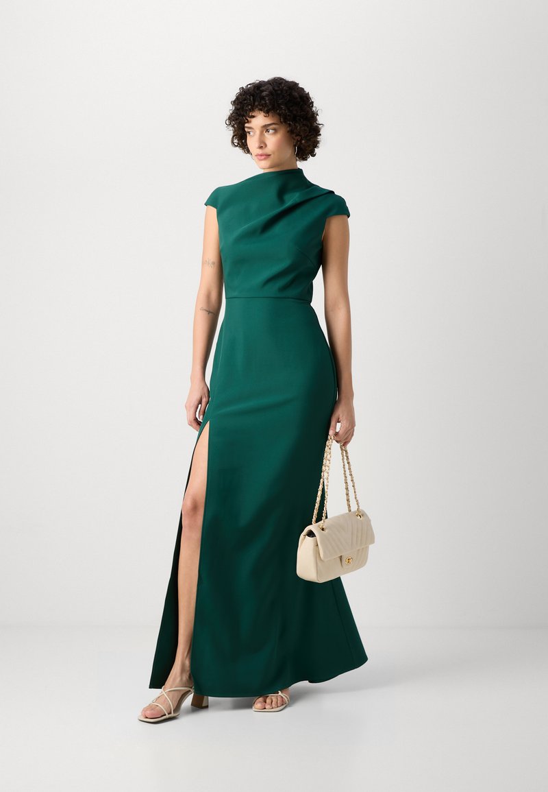Zielona, długa sukienka z wysokim dekoltem i bocznym rozcięciem. Gładki materiał, krótkie rękawki, w połączeniu z beżową torebką i sandałami na paski.