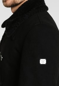 Manteau noir avec un col doux et texturé, arborant un patch logo argenté sur la manche et des fermetures à boutons ronds à l'avant.