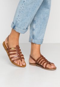 Sandali in pelle marrone con più cinturini, punta aperta, suola piatta e cinturino alla caviglia regolabile, abbinati a jeans in denim azzurri arrotolati.
