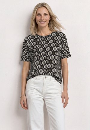 Smilende kvinde med skulderlangt blondt hår iført en sort og beige mønstret kortærmet top og hvide bukser mod en ensfarvet væg.