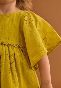 Gelbes Baumwollkleid mit gestickten Blumenmustern, gerafftem Taillenbund und weitem Ärmel. Weicher Stoff mit Spitzelementen.