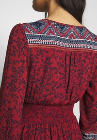 Femme portant une robe rouge avec des motifs floraux bleu marine et un empiècement à motifs géométriques, vue de dos, de la taille vers le haut.
