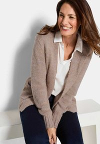 Frau sitzt auf einer Bank, trägt eine beige Strickjacke, ein weißes Hemd mit Kragen und dunkle Hosen, lächelt und schaut zur Seite.