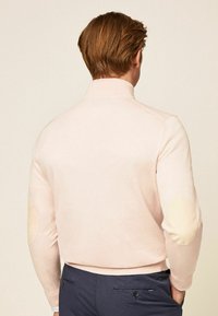 Světle růžový pulovr s vysokým límcem, žebrovaným lemem a loketními záplatami v kontrastní hnědé barvě. Hladká textura, přiléhavý střih.