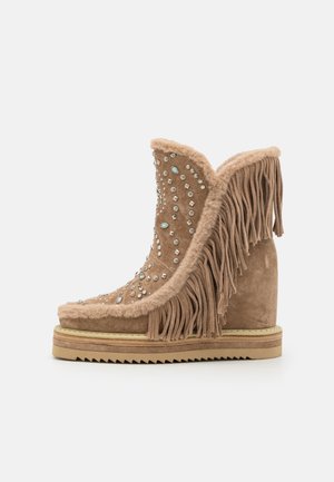 Bottine en suede beige à la cheville avec des détails en franges et ornée de strass, dotée d'un talon en wedge et d'une semelle texturée.