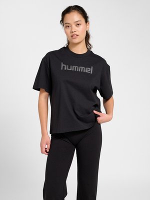 Ung kvinde står med den ene hånd i lommen, iført en løs sort "hummel" t-shirt og sorte bukser mod en ensfarvet baggrund.