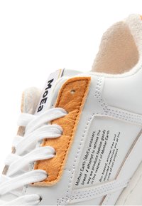 White sneakers with orange felt accent, textured tongue, and laces. Side label with product description in black text on white background.