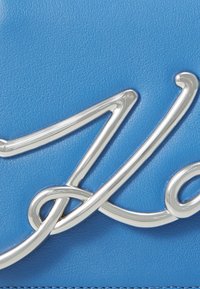 Borsa a tracolla in pelle blu con un'inserzione di logo in metallo argentato lucido, caratterizzata da una consistenza liscia e un design elegante e fluido.