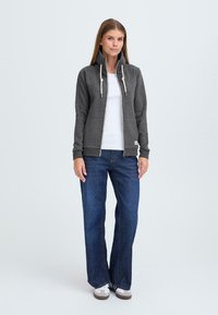Grijze zip-up hoodie met hoge kraag, trekkoorden, zijzakken, ribgebreide manchetten, gecombineerd met blauwe wijde jeans en witte sneakers.