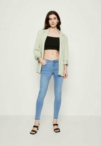 Ανοιχτό πράσινο oversized σακάκι πάνω από ένα μαύρο crop top, συνδυασμένο με μπλε skinny jeans και μαύρες ψηλές σαγιονάρες. Λείες υφές, χαλαρή εφαρμογή.