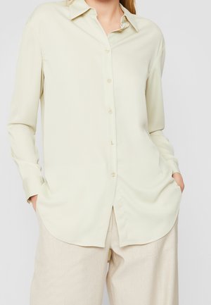 Femme portant une chemise beige clair à manches longues boutonnée avec col et un pantalon ample couleur crème, mains dans les poches.