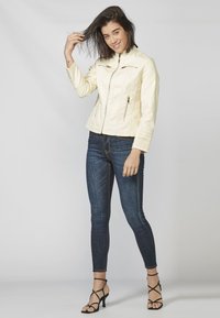 Giacca attillata color crema con tasche con cerniera e accenti ricamati, abbinata a jeans skinny blu scuro e tacchi neri con cinturini.
