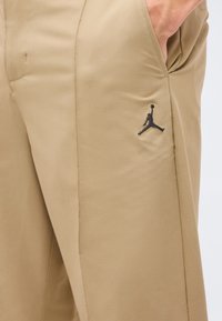 Κοντινή λήψη μπεζ αθλητικών παντελονιών με χέρι στην τσέπη και μικρό μαύρο λογότυπο Jumpman κεντημένο στον μηρό.