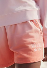 Roze sweatshirt met geribbelde manchetten en lichtroze ombre-shorts, voorzien van de borduursel "MIAMI Est 1987" in het wit. Zachte, gladde stoftextuur.
