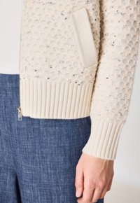 Cardigan en tricot couleur crème avec un motif texturé, des bords côtelés aux poignets et au bas, et une poche latérale. Fermeture éclair avec quincaillerie dorée.