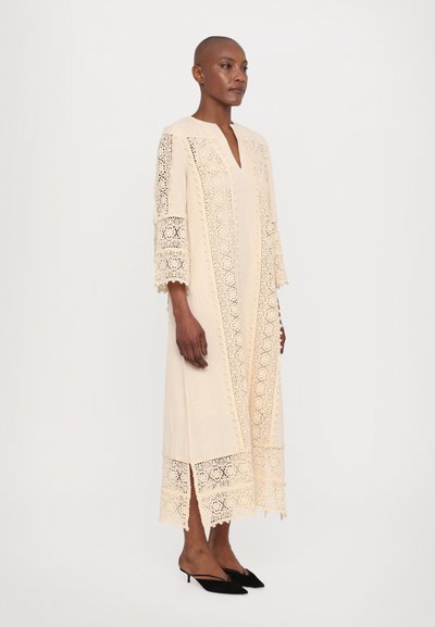 Beige dress made of lace with intricate cutout patterns, features a V-neck, 3/4 sleeves, and side slits for ease of movement.