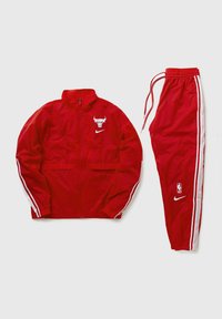 Giacca a vento rossa e pantaloni coordinati, entrambi realizzati in materiale leggero. Presentano strisce laterali bianche e loghi. Chiusura con zip davanti, polsini a costine.