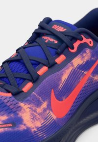 Μπλε και μοβ αθλητικό παπούτσι με υφασμάτινο επάνω μέρος, πορτοκαλί λεπτομέρειες και εμφανές λογότυπο Nike. Διαθέτει δέσιμο με κορδόνια και μαλακή σόλα.