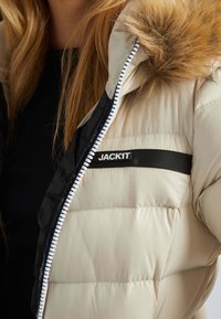 Pufferjack in lichtbeige met een faux fur capuchon, zwarte binnenkant met een textuurpatroon, en een witte rits. Logo op de zak in zwart.