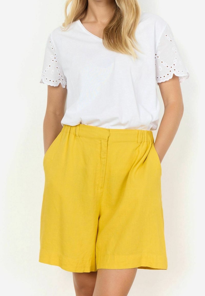 Femme portant un haut blanc à manches courtes avec des détails œillets et un short taille haute jaune vif avec des poches latérales, mains dans les poches.