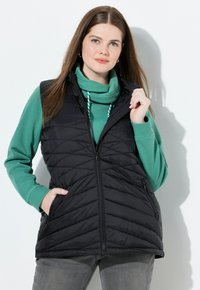 Schwarze Steppweste mit einem Reißverschluss, gestaltet mit einem hohen Kragen und zwei Seitentaschen, getragen über einem grünen Sweatshirt.