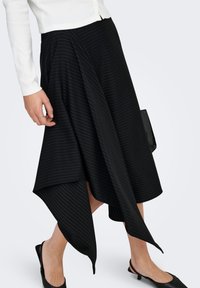 Jupe asymétrique à rayures noires avec un design fluide, confectionnée en tissu texturé, portée avec un haut blanc et des mules noires à talons.