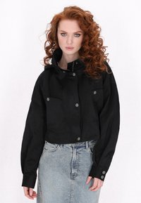Jachetă neagră scurtă, realizată dintr-un material neted, cu un guler înalt, închidere cu butoane pe față și două buzunare pe piept. Combinată cu o fustă din denim.