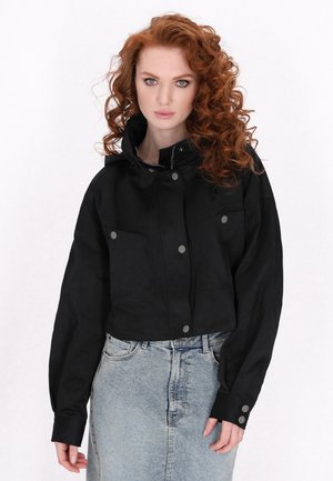 Black cropped jacket made of a smooth fabric, featuring a high collar, front button closures, and two chest pockets. Paired with a denim skirt.
