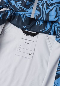 Blue and black patterned jacket with a soft textile lining, featuring a care tag and hanging loop. Sustainable design noted.