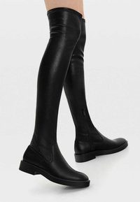 Botas de cuero negras hasta la rodilla con punta redonda, suela plana y cremallera lateral. Textura suave y diseño ajustado.