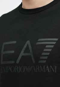 Közeli kép egy fekete Emporio Armani EA7 pólóról sötétszürke logóval és felirattal a mellkasán.