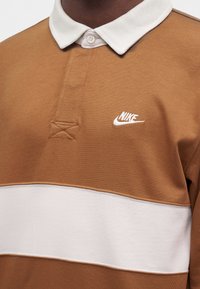 Καφέ και λευκό πόλο Nike με λευκό γιακά και κεντημένο λογότυπο Nike στο στήθος.