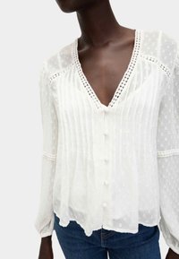 Blusa bianca con scollatura a V profonda, tessuto testurizzato con strisce e pois alternati e chiusura con bottoni. Maniche ampie con polsini arricciati.
