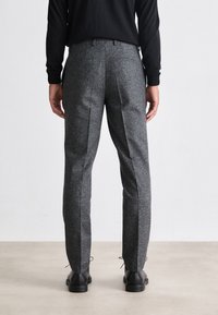 Pantaloni grigi, texture leggermente lucida, con vestibilità su misura e tasche posteriori, abbinati a scarpe nere e una camicia a maniche lunghe.