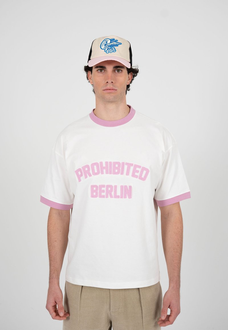 Mann trägt beige Hose, weißes T-Shirt mit pinker Aufschrift "PROHIBITED BERLIN" und beige-pinke Kappe mit blauem Grafikdruck vor weißem Hintergrund.
