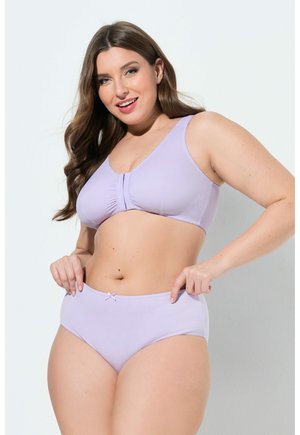 Uśmiechnięta kobieta w jasnofioletowym komplecie bielizny, regulująca pas, stojąca na jednolitym białym tle.