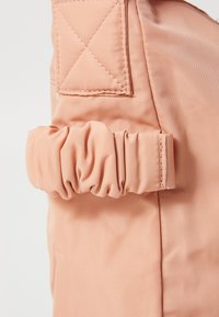 Zachte perzikkleurige stoffen tas met geraffineerde elastische details, vierkant label met kruissteekaccenten, gladde textuur en minimalistisch ontwerp.