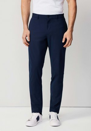 Pantalons slim-fit bleu marine en tissu léger, avec une fermeture à bouton sur le devant, des poches latérales, et associés à des baskets blanches.