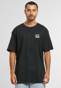 Black cotton t-shirt with short sleeves, featuring a small green and white logo on the left chest area, relaxed fit, round neckline.