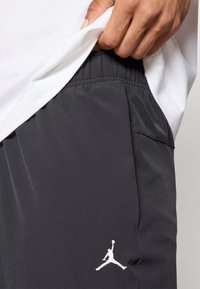 Μαύρο αθλητικό σορτς με λεία υφή, ελαστική μέση και λευκό λογότυπο Jumpman στη αριστερή πλευρά. Ένα λευκό T-shirt είναι ελαφρώς μισο tucked.