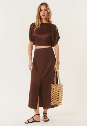 Jeune femme portant un top court marron et une jupe assortie avec une fente devant, des sandales noires, des bracelets dorés et un sac à main beige tissé.