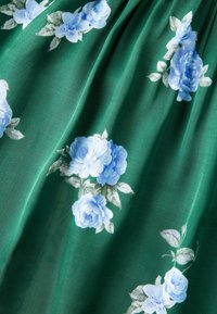 Tissu vert avec un motif répétitif de roses bleu clair et de feuilles vert pâle. Le tissu présente de doux plis et une texture lisse.