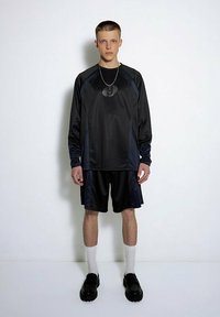 Schwarzes Sport-Outfit bestehend aus einem Langarmshirt und Shorts mit Struktur. Die Accessoires umfassen eine kreisförmige Anhänger-Halskette.